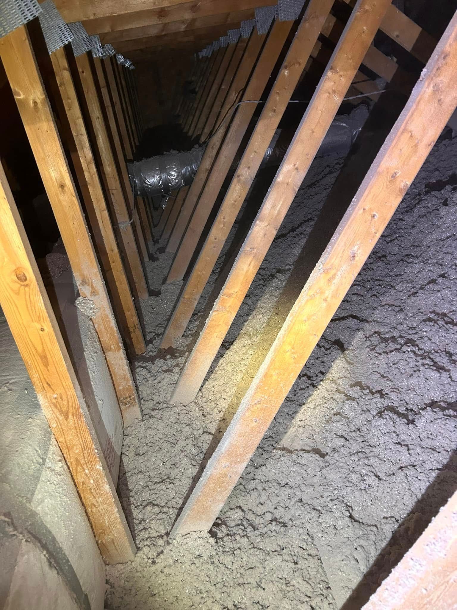Inside a dusty attic. Wood beams form a V-shape. Loose, light-colored insulation fills the space.
