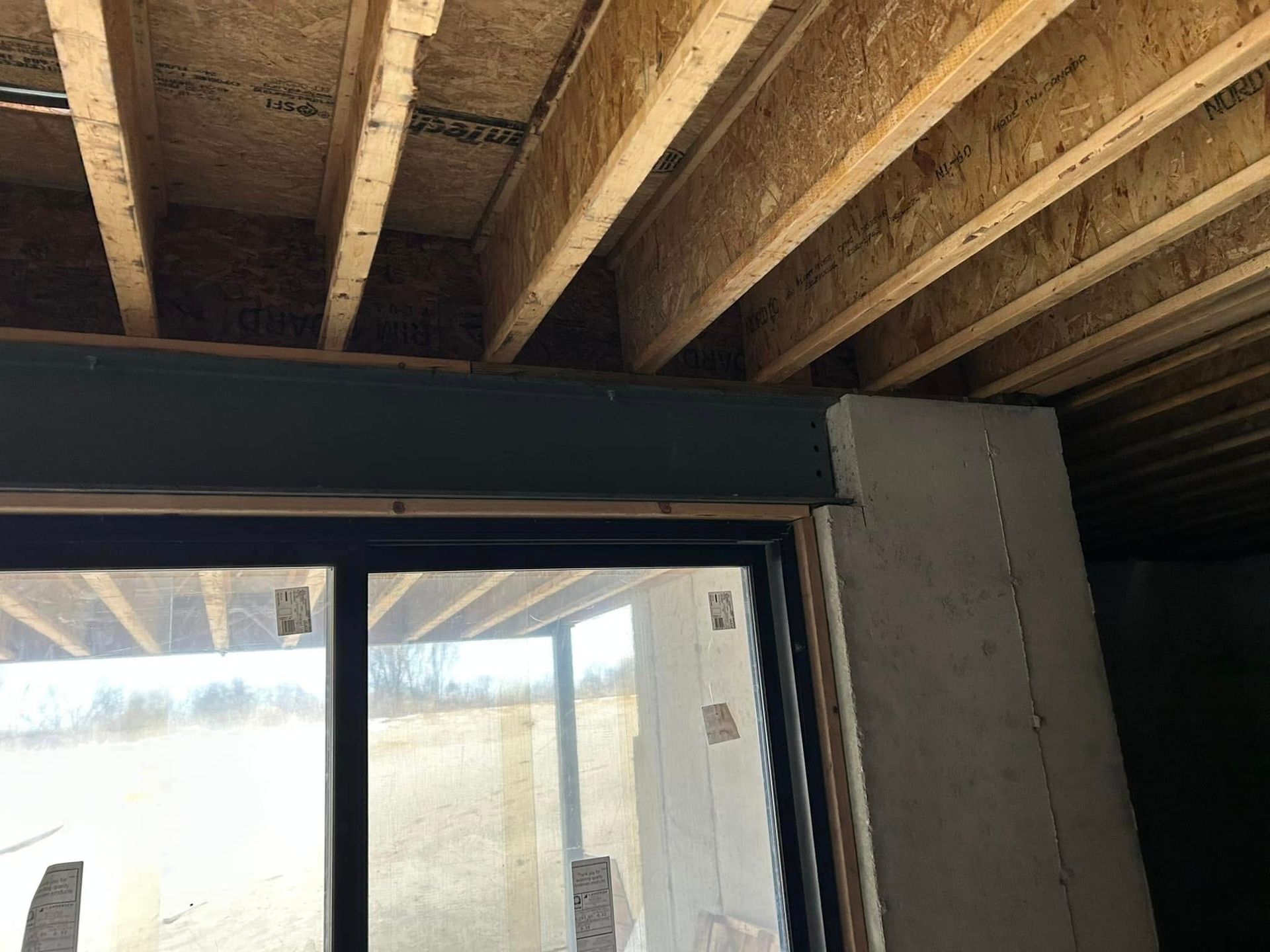Interior view of a building under construction, showing a window, dark beam, wood joists, and concrete column.