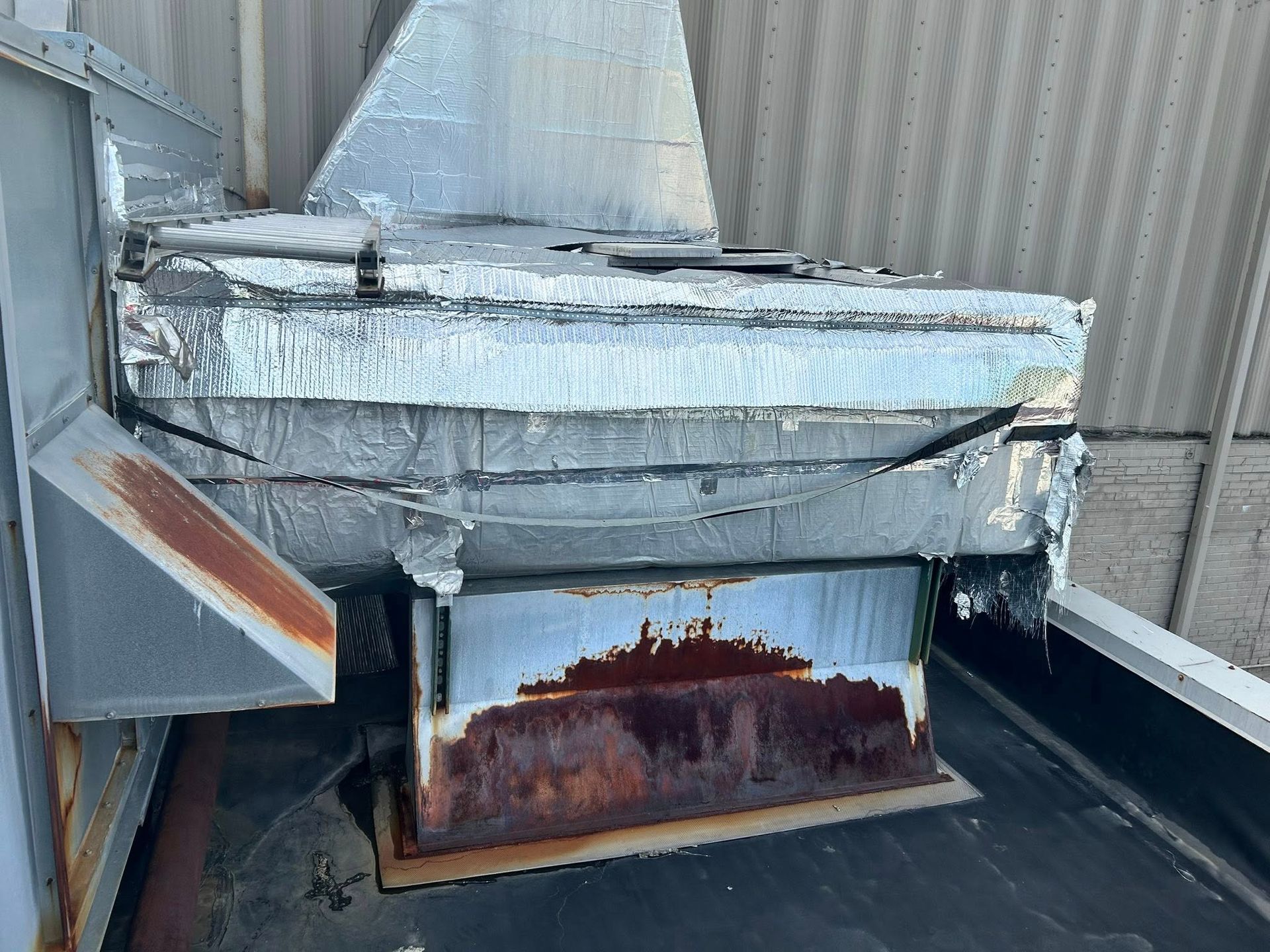 Rusted rooftop HVAC unit covered in insulation and reflective tape, with a dark roof background.