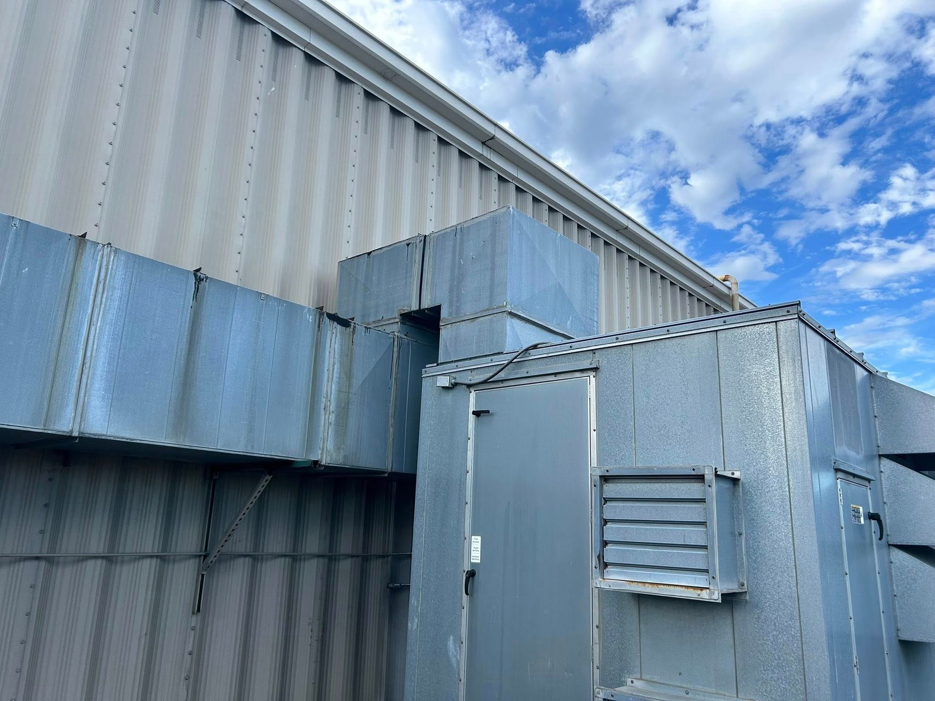 Exterior view of a metal building with silver ventilation ducts, a door, and a window; cloudy sky in the background.