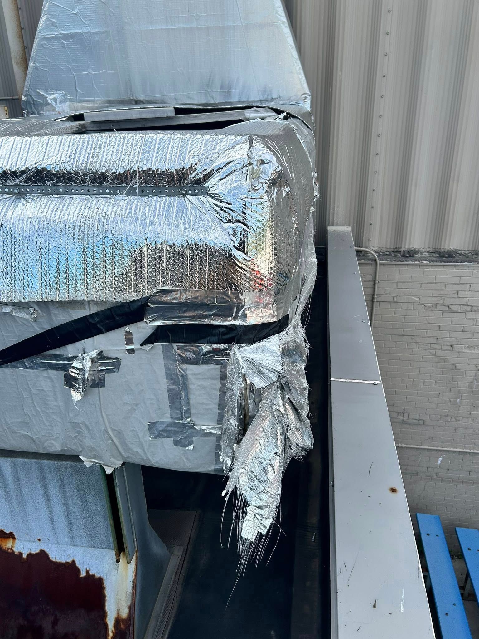 Large, insulated metal box, wrapped in silver foil, partially resting on a metal shelf near a building.