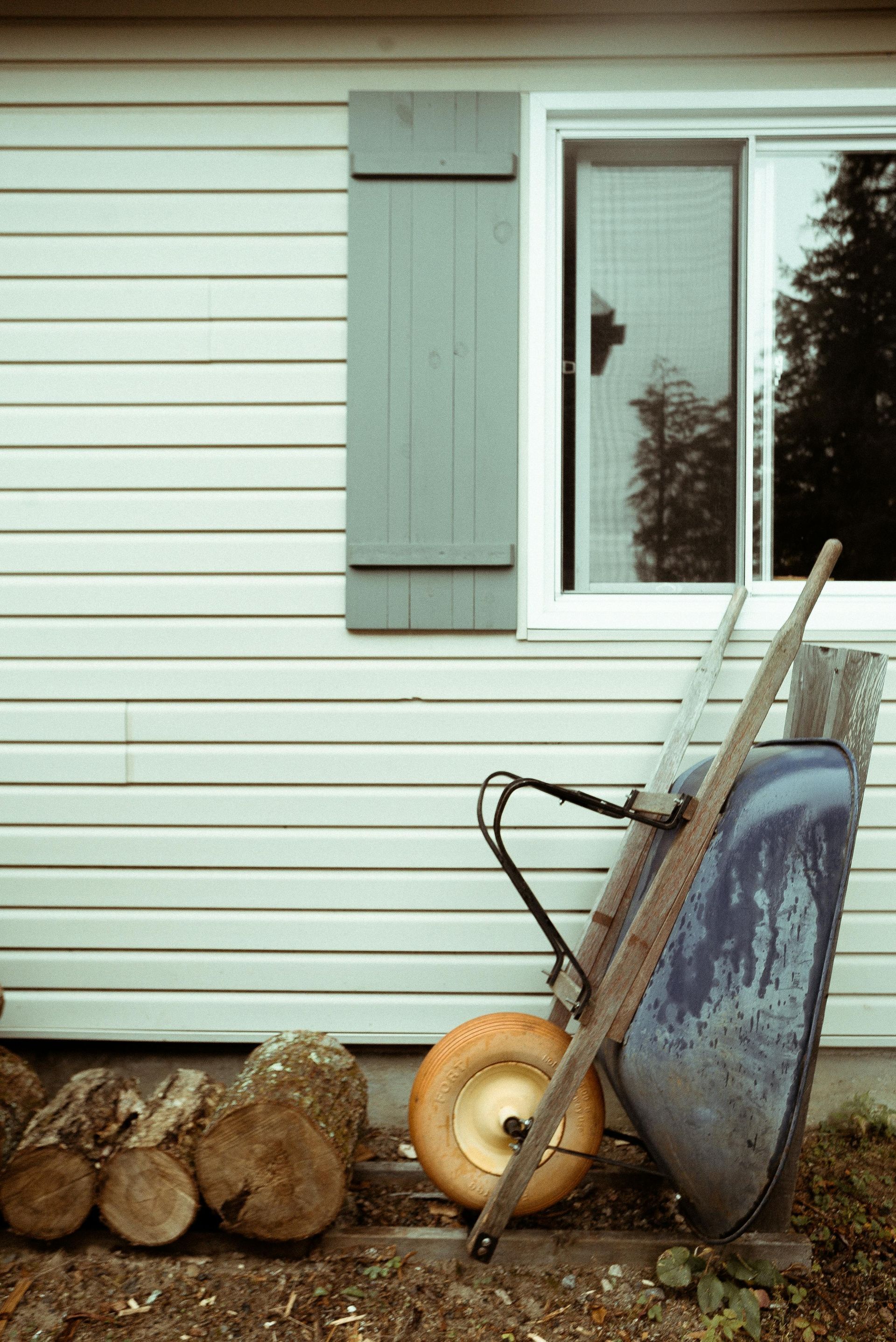 A wheelbarrow is sitting in front of a house next to a pile of logs.
