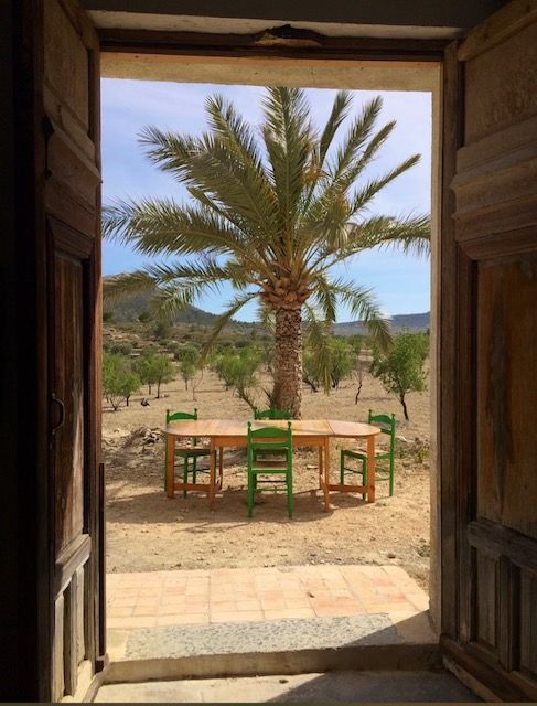 Een open houten deuropening biedt uitzicht op een woestijnlandschap met een tafel, stoelen, een palmboom en heuvels.