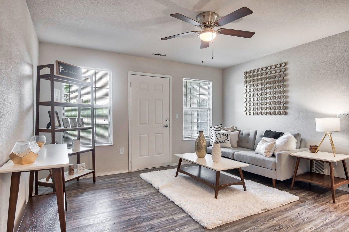 Living room with gray sofa, coffee table, and rug. Windows, shelving unit, and art are also present.