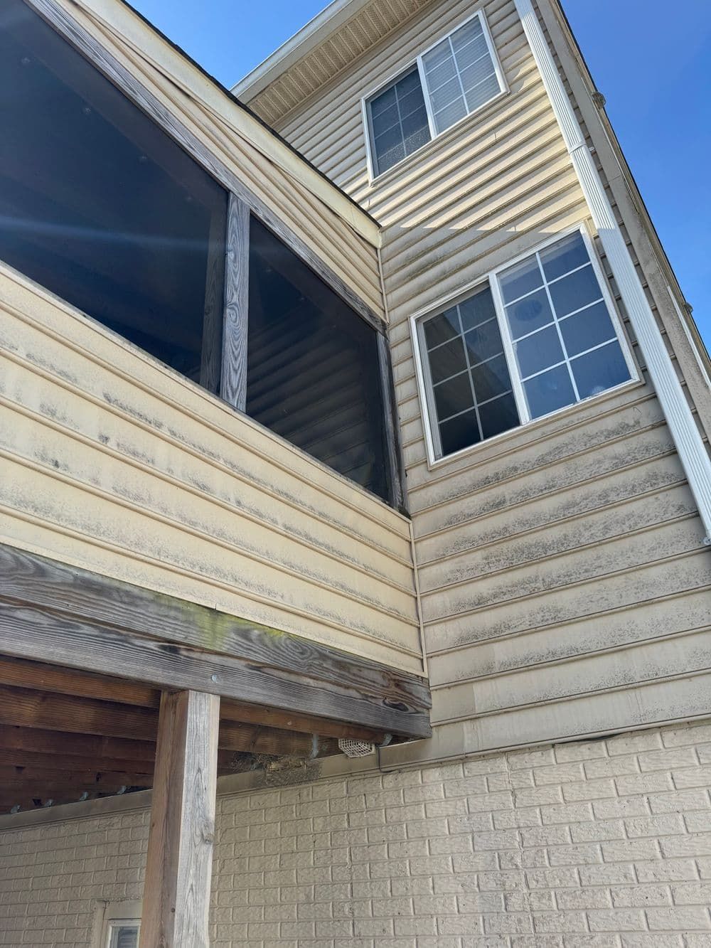 Beige siding of a two-story house, with a screened porch and two white-framed windows against a blue sky.