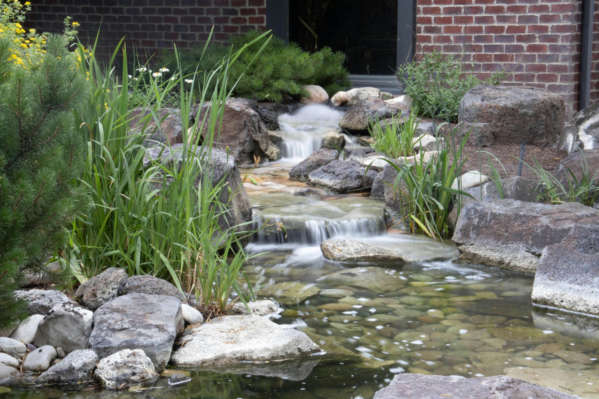 Rock garden water feature with a small stream cascading over stones beside green plants and a brick wall
