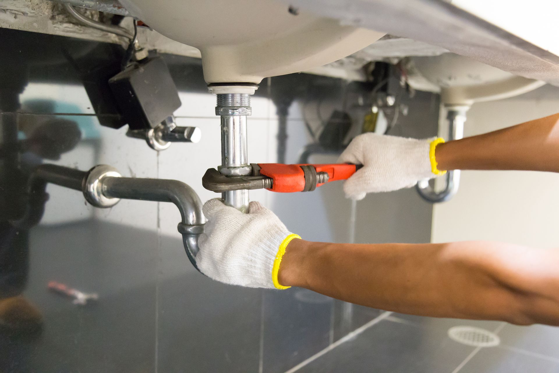 A person using a wrench to work on plumbing under a sink. They are wearing gloves.