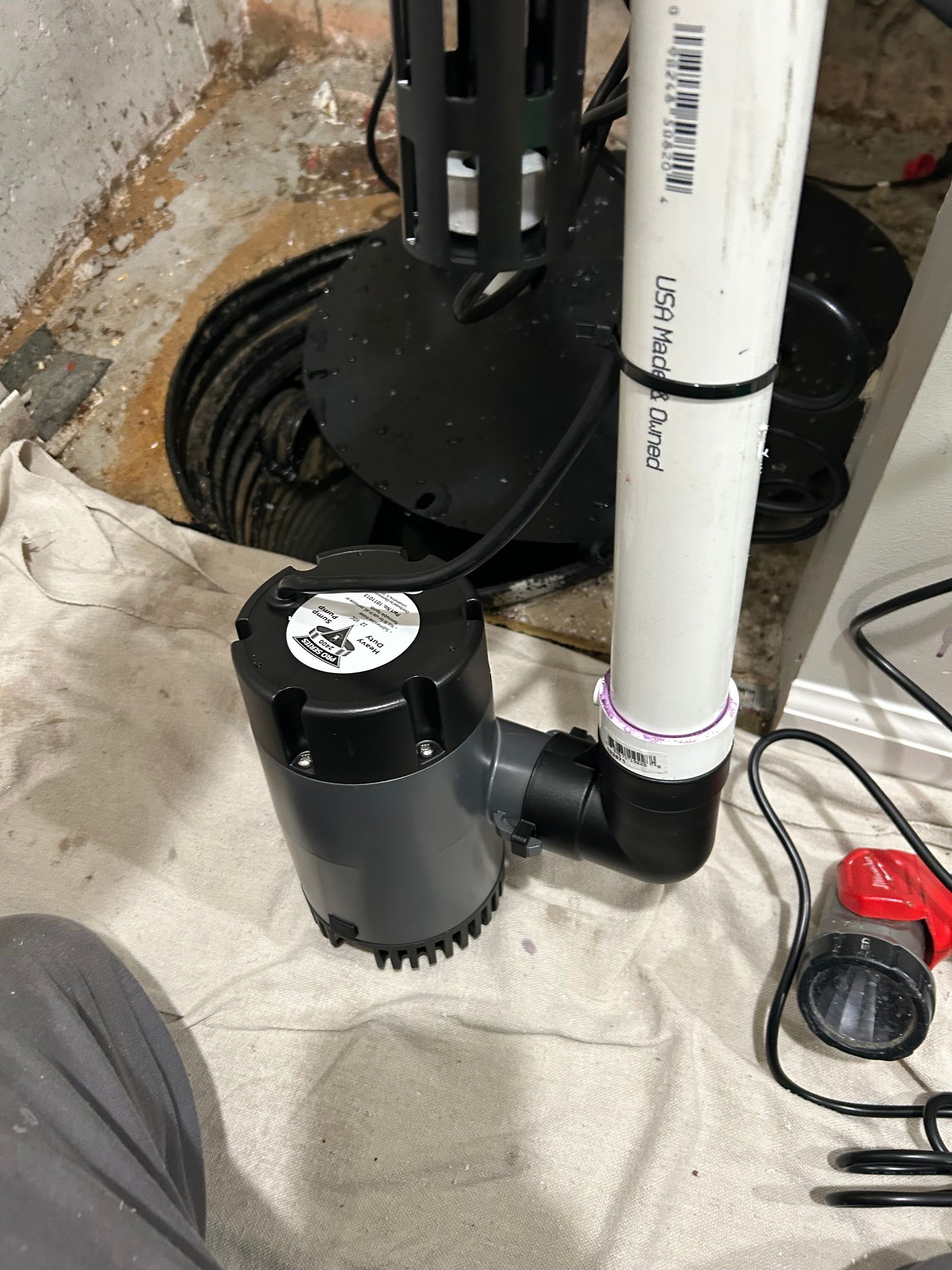 Person operating a red and black water pump in muddy water, wearing boots.