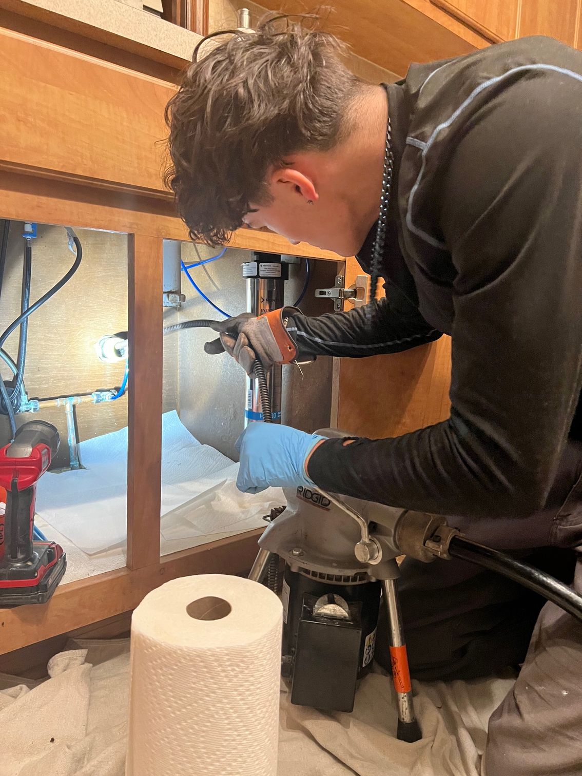 Person using a plumbing snake to unclog a white sink. Hands wear blue gloves. Red and black snake tool.