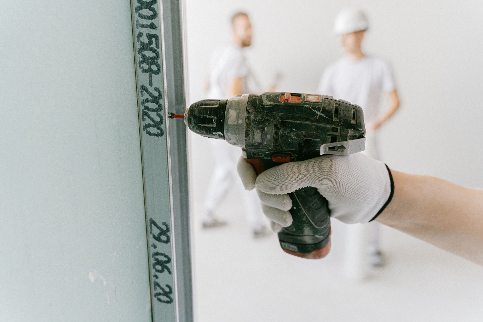 Gloved hand using a power drill on a wall-mounted pipe in a white room with blurred workers in the background