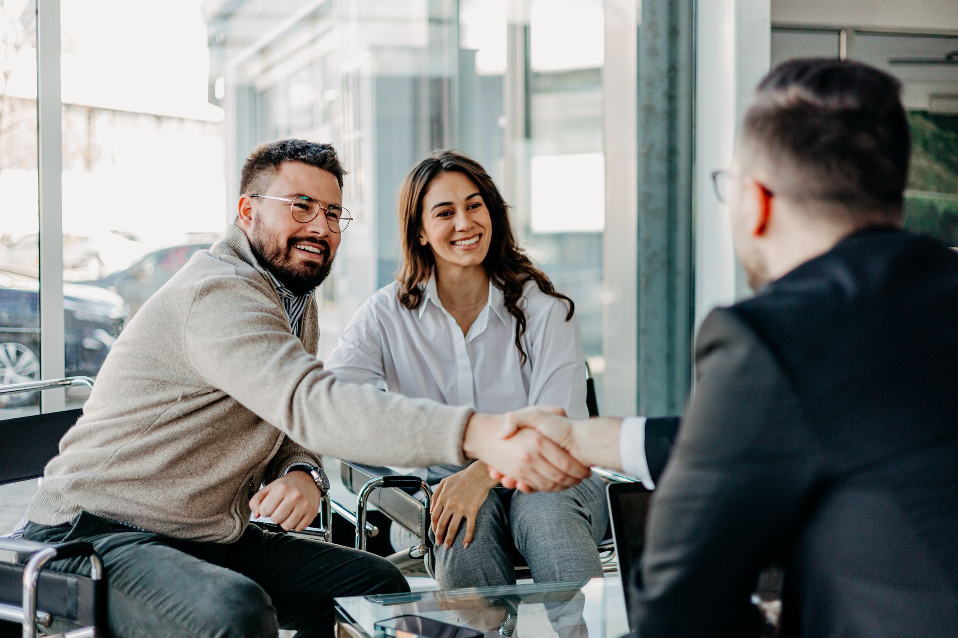 Two people shaking hands in a bright office meeting room, with a third person seated nearby.