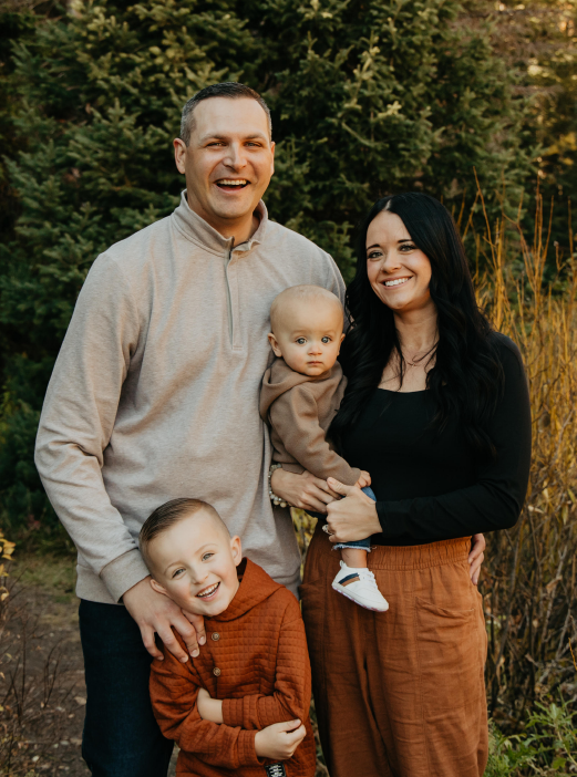 Family of four outdoors, smiling and posing in front of trees; one child is held, another stands in front.