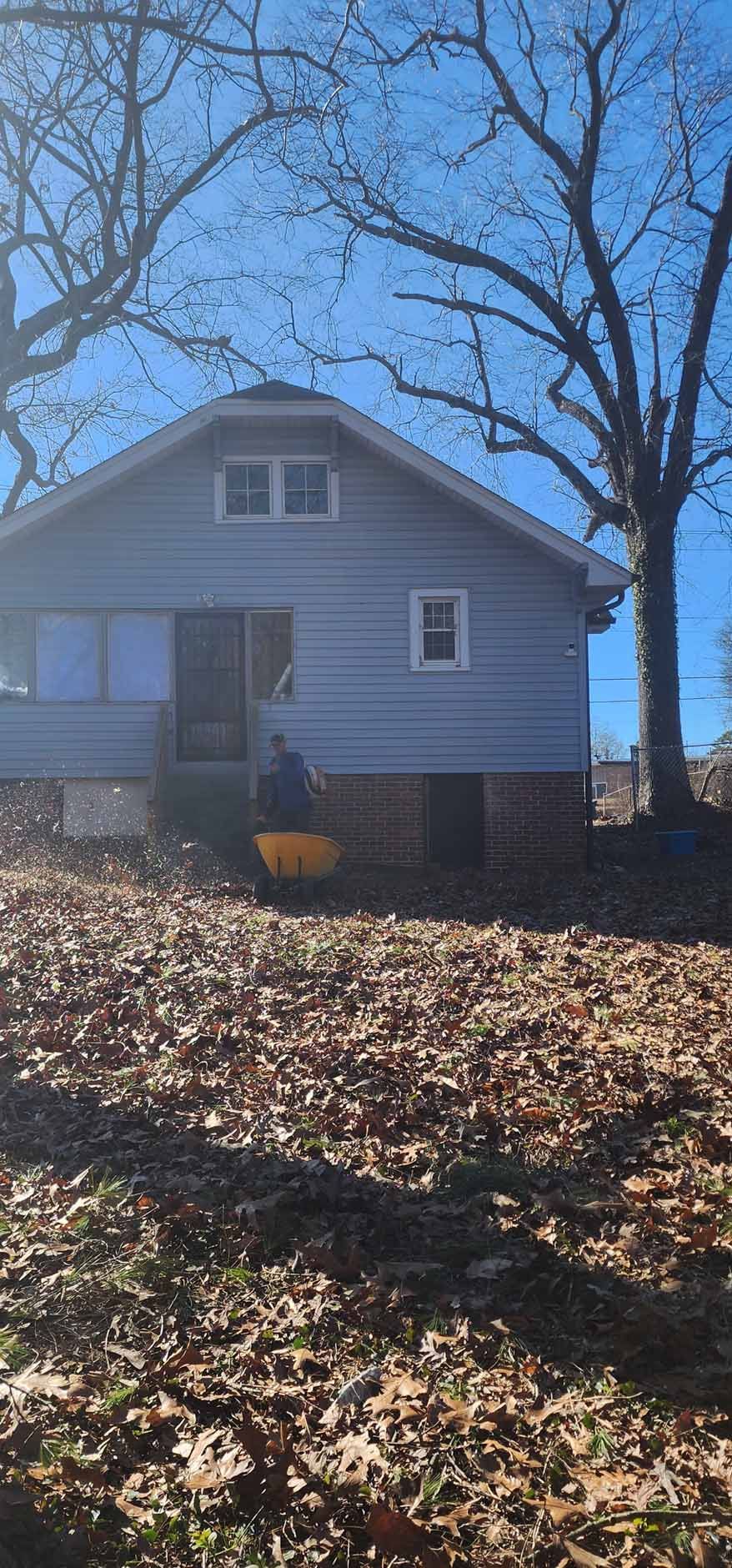 Blue house with a person operating a yellow wheelbarrow; leaves on the ground.
