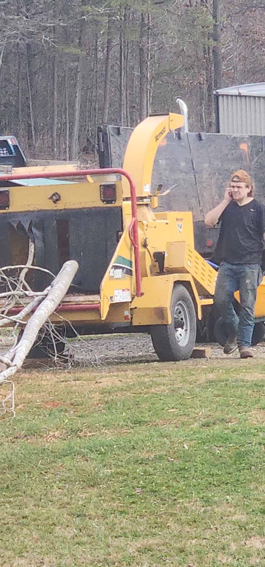A man stands by a yellow wood chipper, talking on a phone. The scene is outdoors with trees in the background.