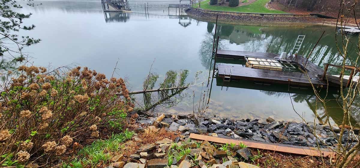 A lake view with a dock and a foggy sky. Dry bushes in the foreground, calm water, and some structures.