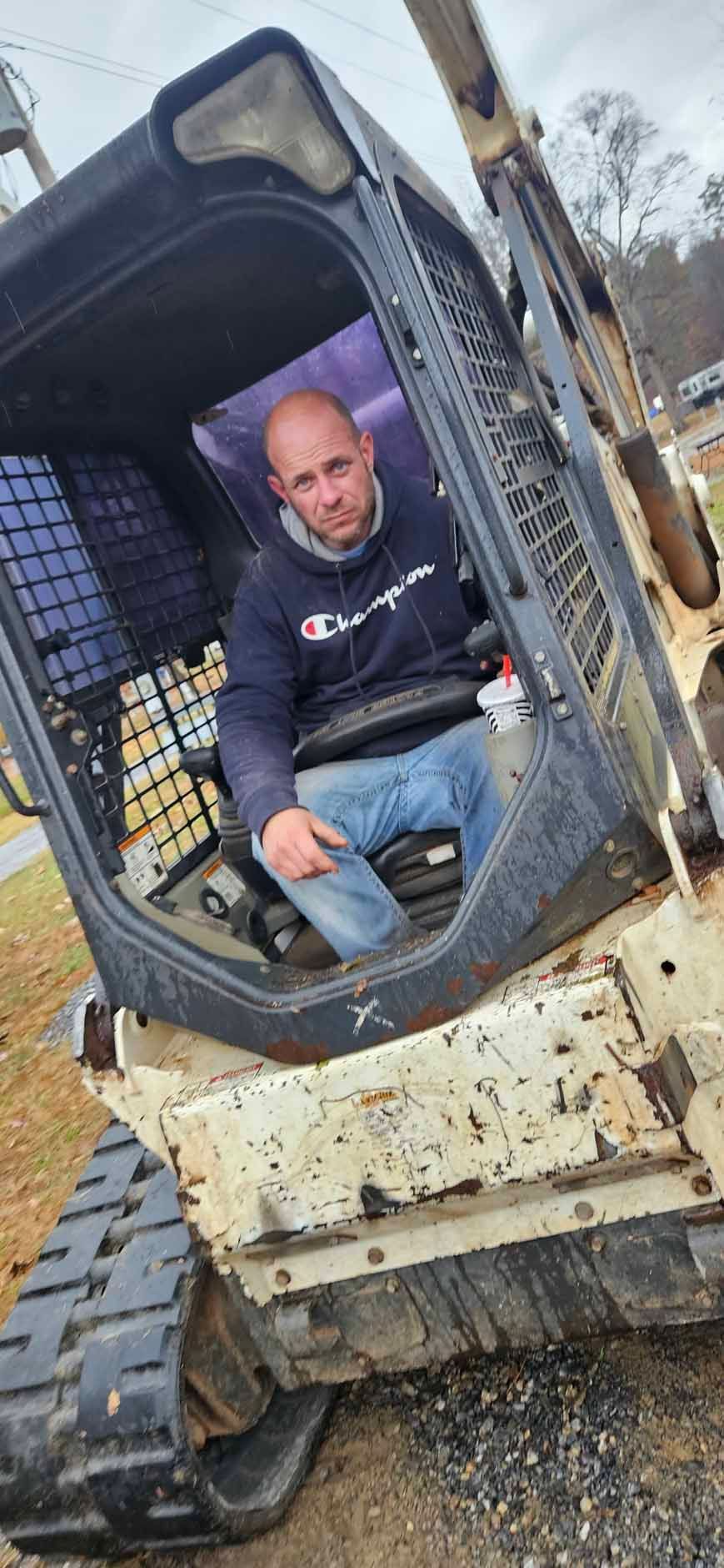 Man in a Champion hoodie inside a skid steer; outdoors.