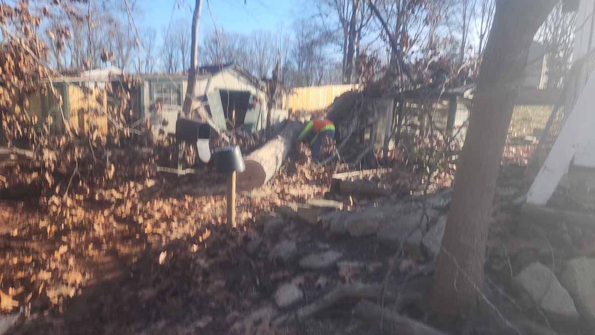 A person in an orange vest is working among debris in a yard with a partially visible house in the background.