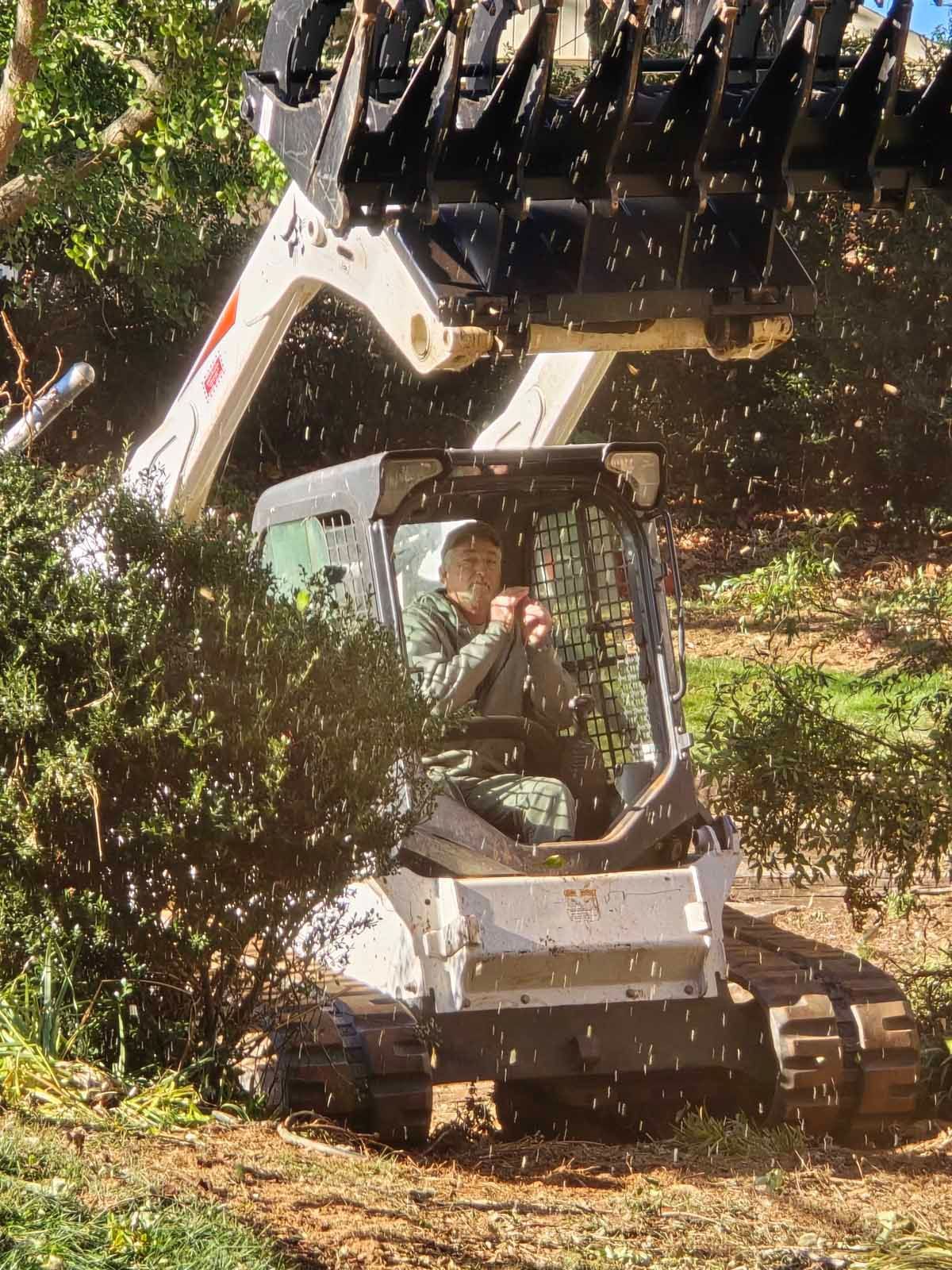 Man operating a white Bobcat clearing brush in a wooded area; debris flying.
