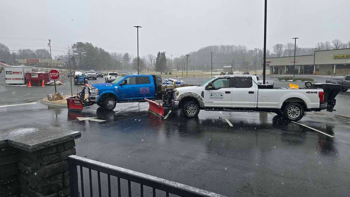 Two snow plows clearing a parking lot in the snow; one blue, one white.