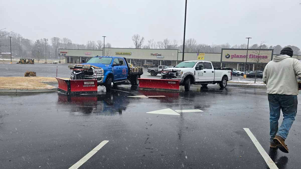 Two snowplow trucks in a wet parking lot clearing snow, near a shopping center on a snowy day. A person walks away.