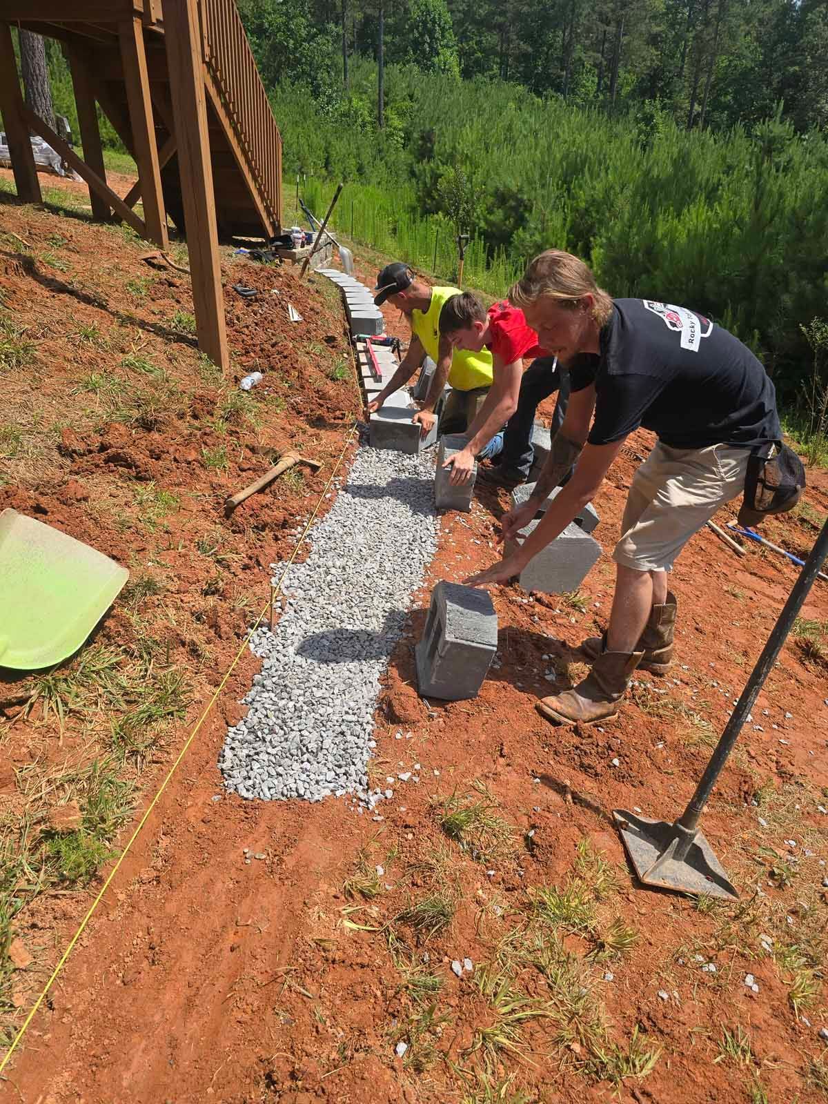 Four workers laying stone blocks to create a retaining wall along a sloped hillside.