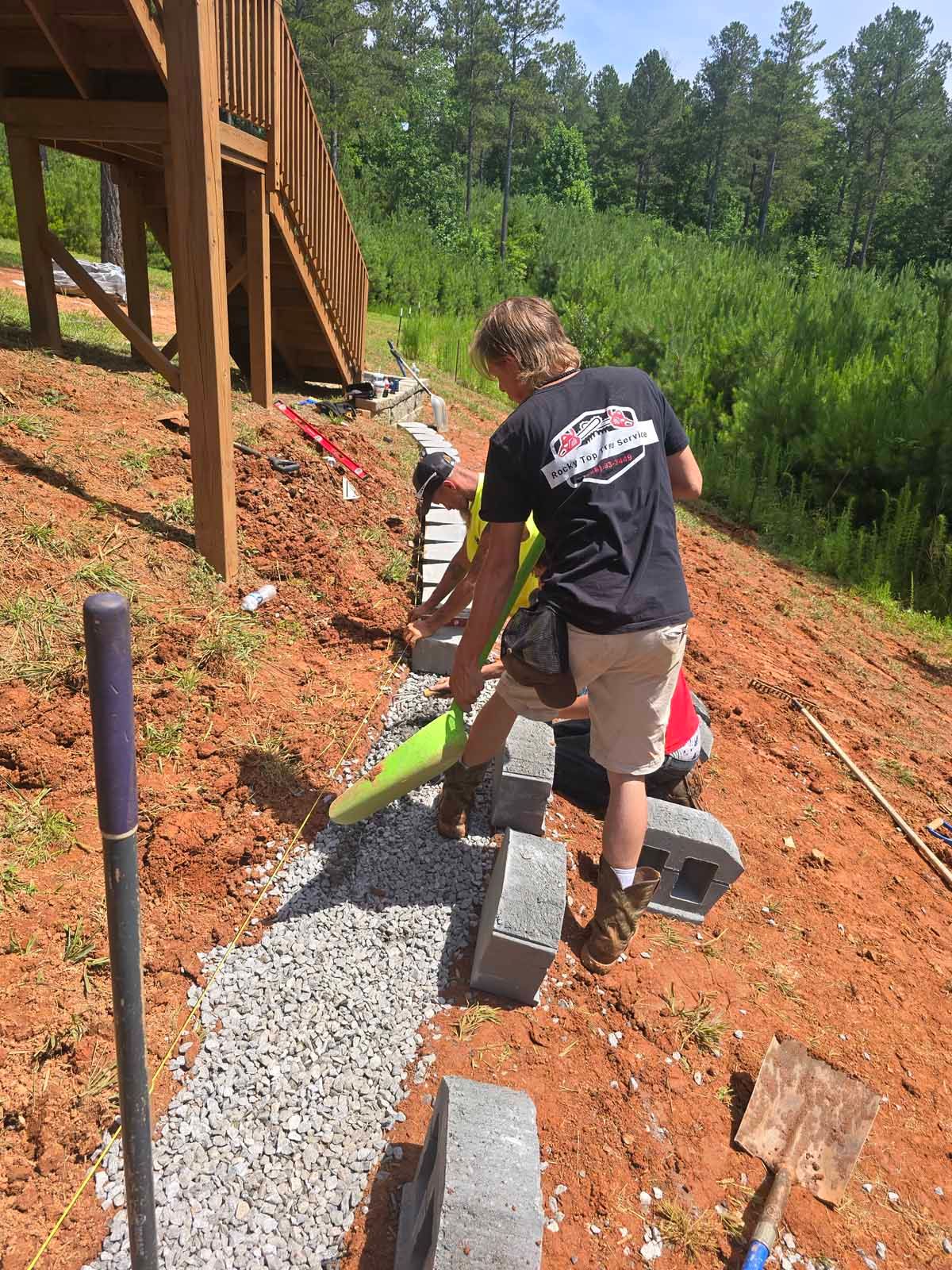 Two men building a retaining wall on a hillside near a wooden deck.