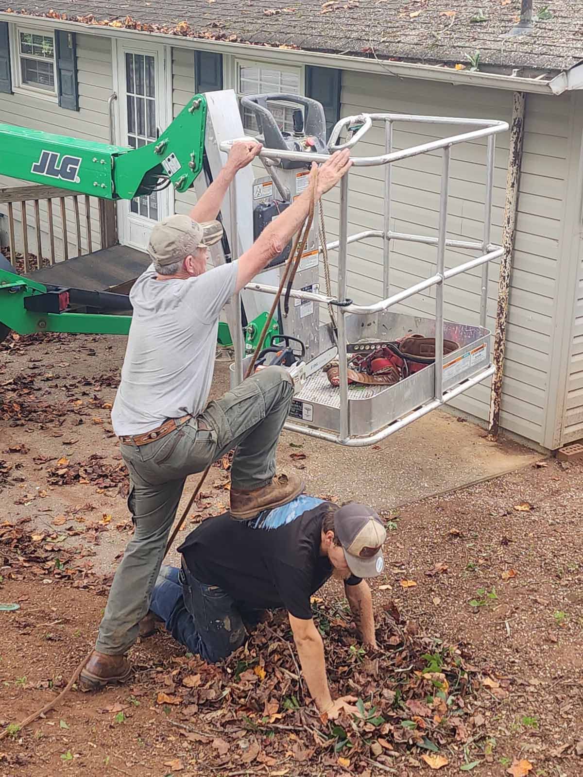 Two men working: one in lift basket, one on the ground, cleaning debris in a yard, with a house in the background.
