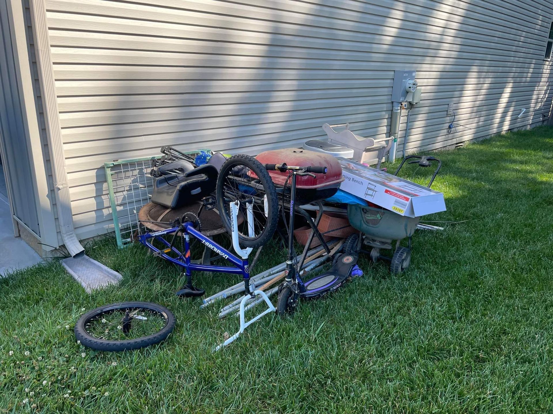 Pile of discarded items on grass next to a light-colored house: bike parts, grill, wheelbarrow, and more.