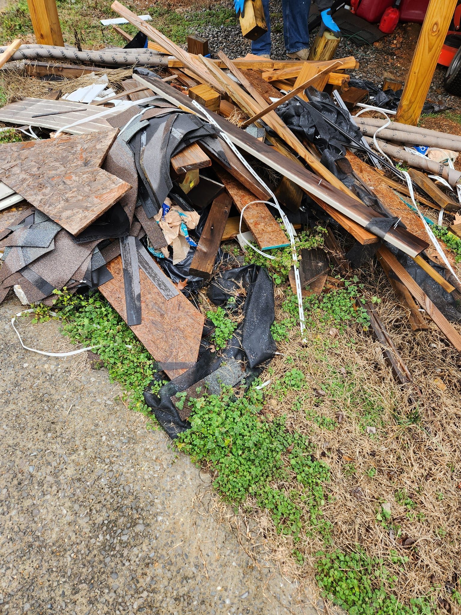 Pile of construction debris: wood, asphalt shingles, and trash on grass next to a concrete area.