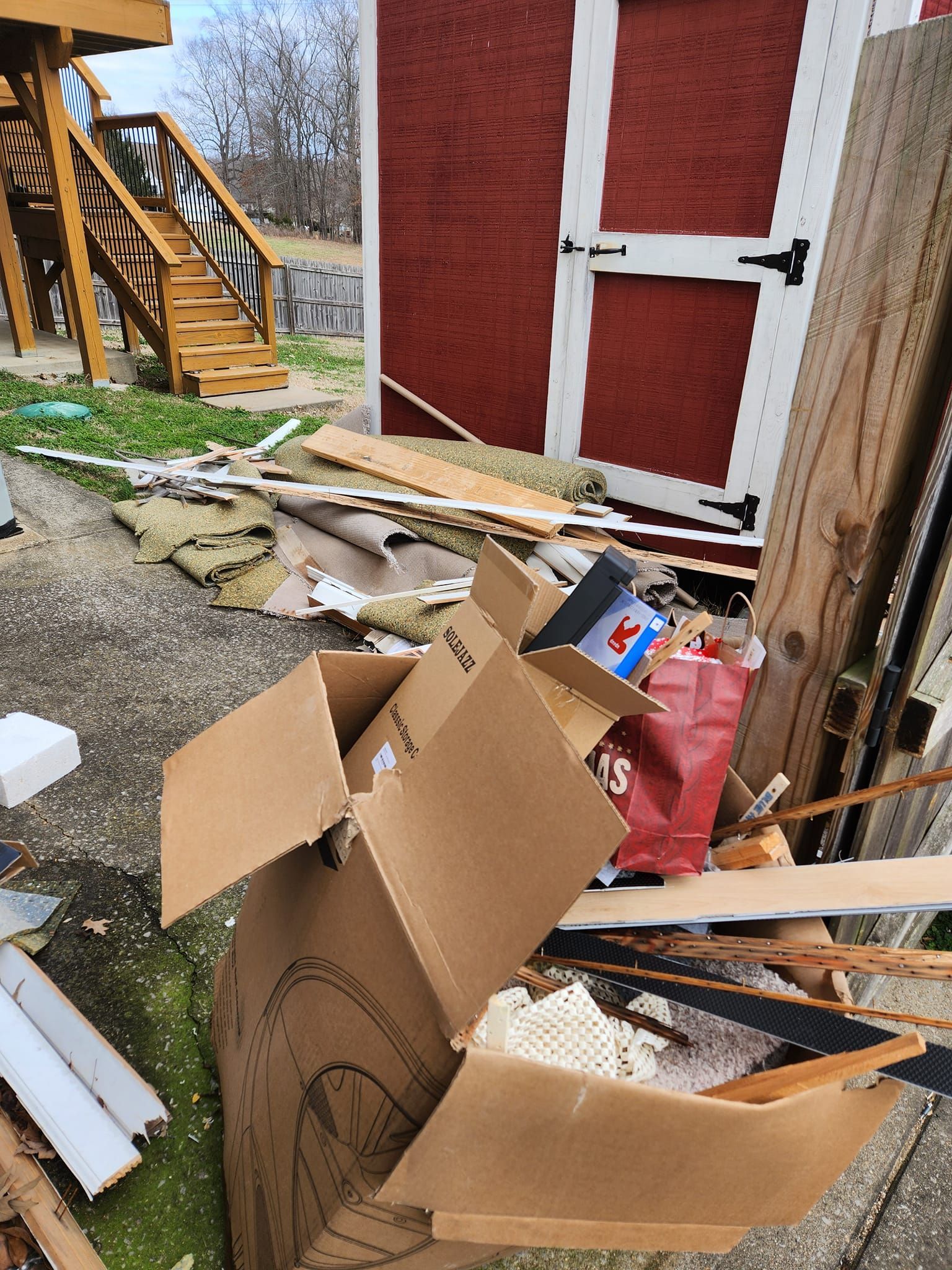 Cardboard box overflowing with trash and debris near a red shed and outdoor stairs.
