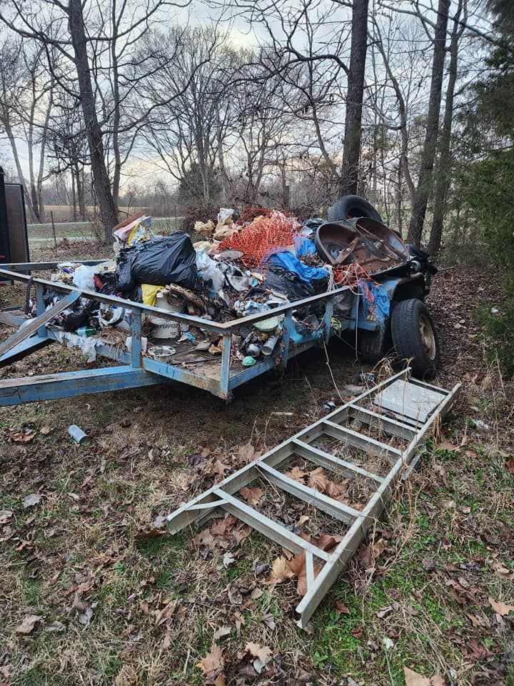 A trailer overflowing with trash and debris in a wooded area, a ladder lies on the ground.