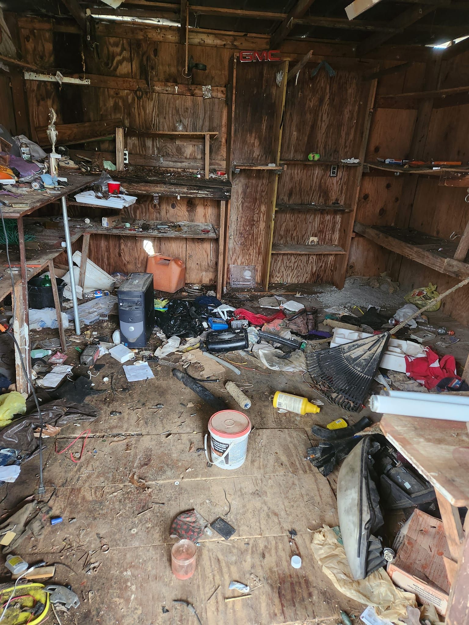 Messy interior of a cluttered, deteriorating wooden shed with scattered items, a paint bucket, and exposed shelving.