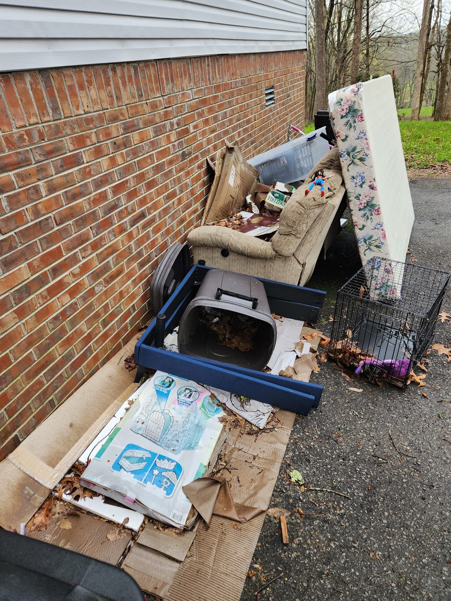 Pile of discarded trash next to a brick wall, including a mattress, chair, and debris on asphalt.