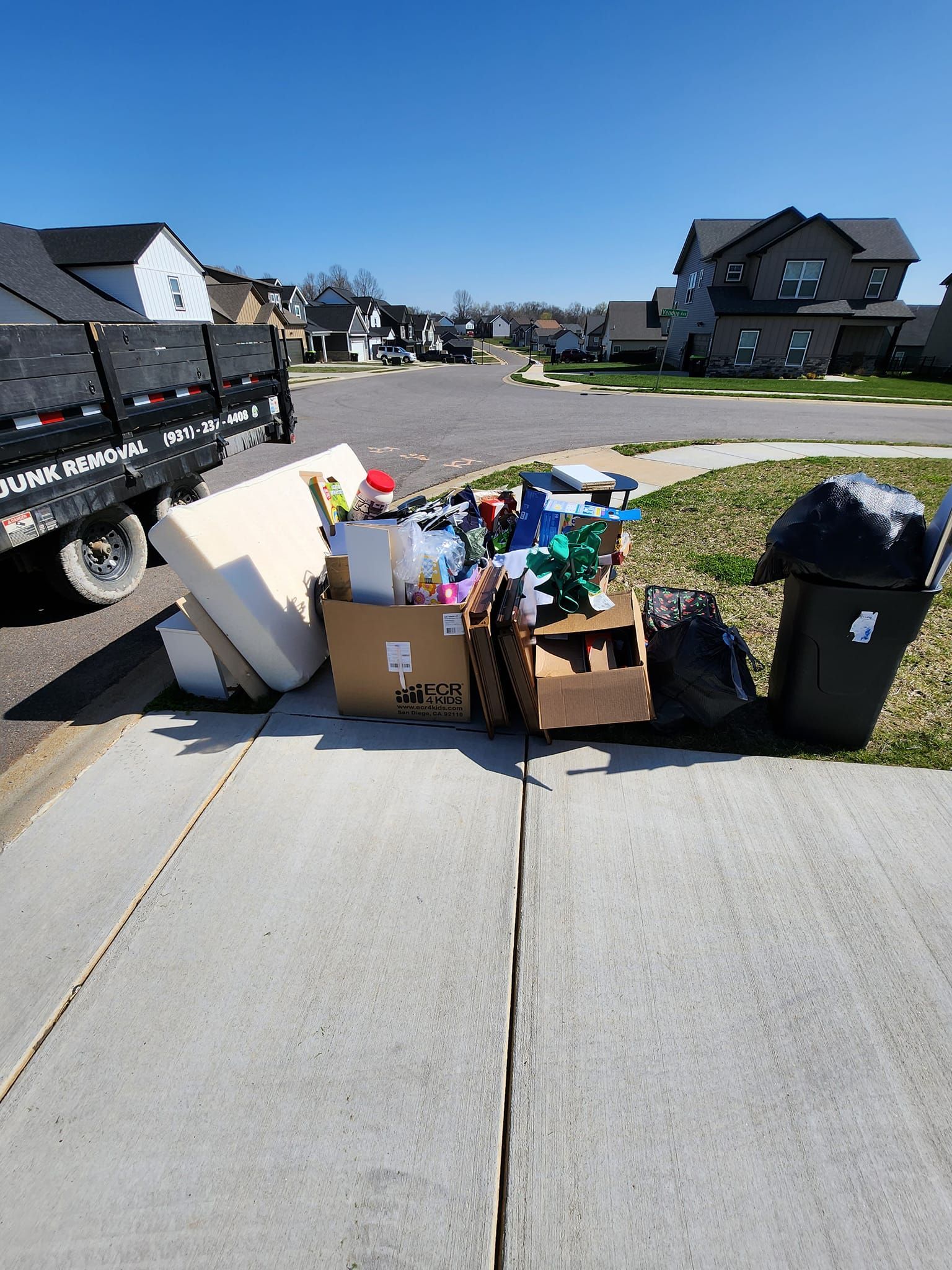 Pile of trash, including a mattress, boxes, and bin, on a sidewalk next to a street and a black trailer.