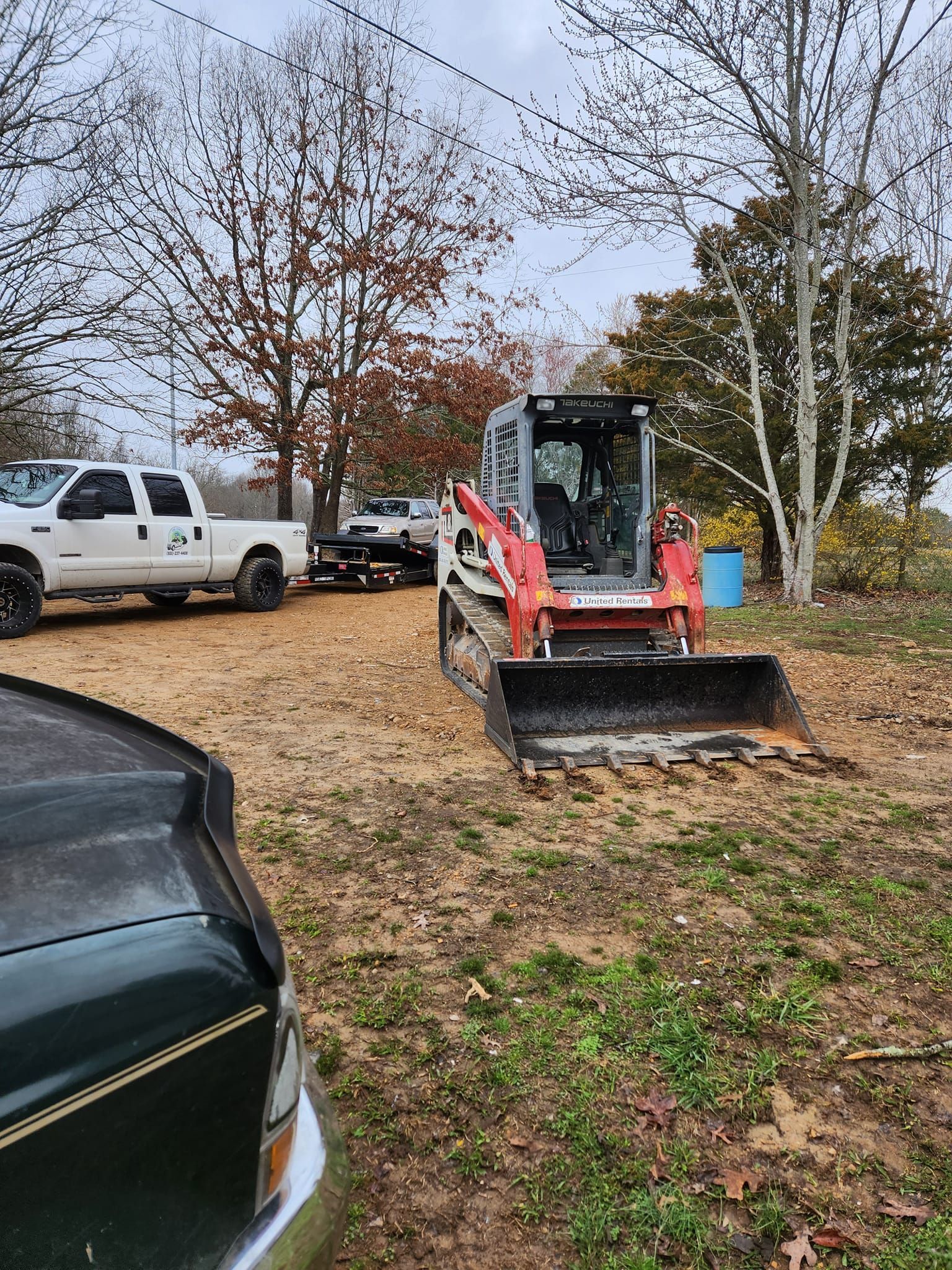 Bobcat in a dirt lot, with truck and trailer in the background. Overcast sky.