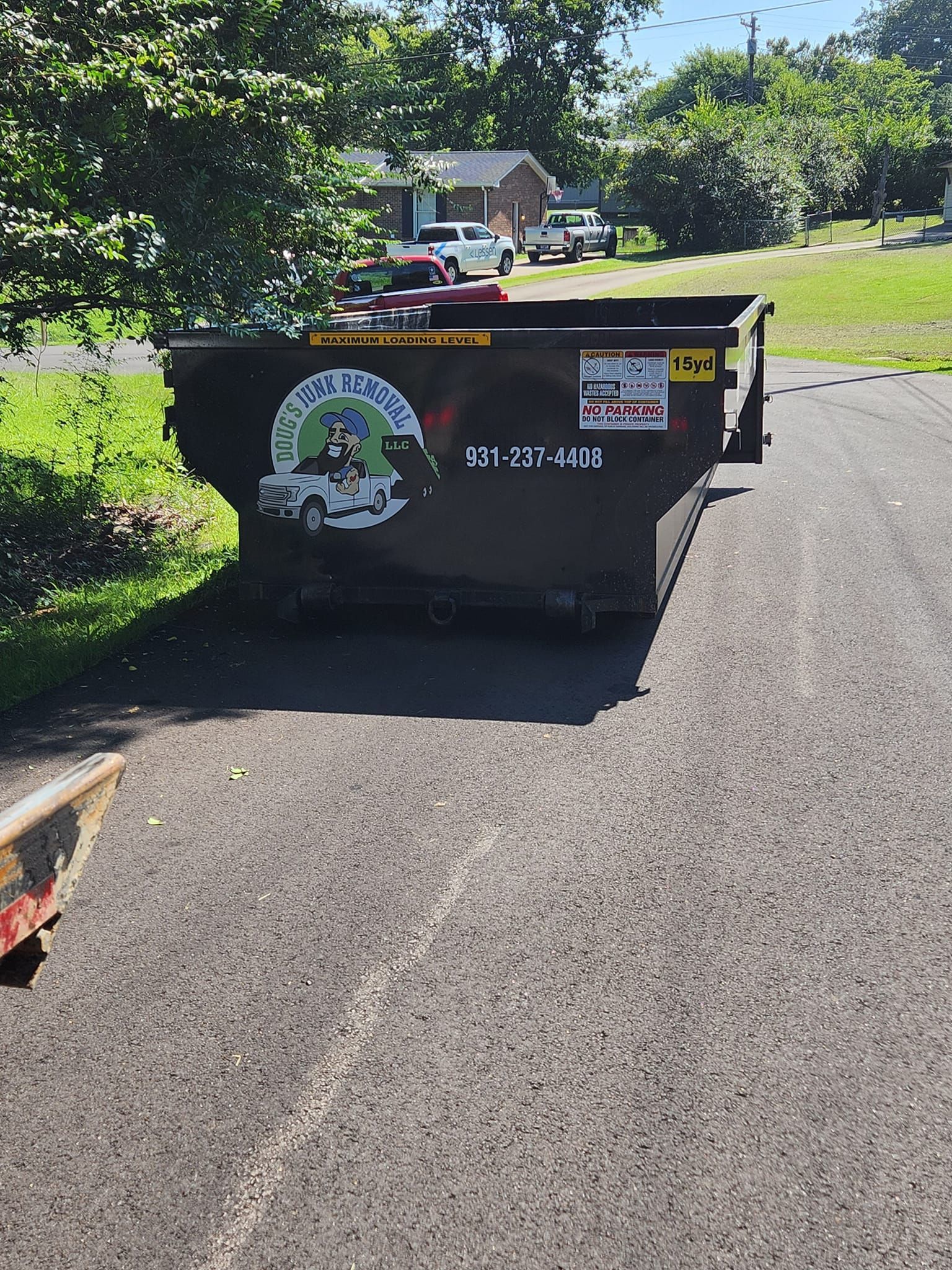 Black dumpster on asphalt driveway with trees and houses in the background.