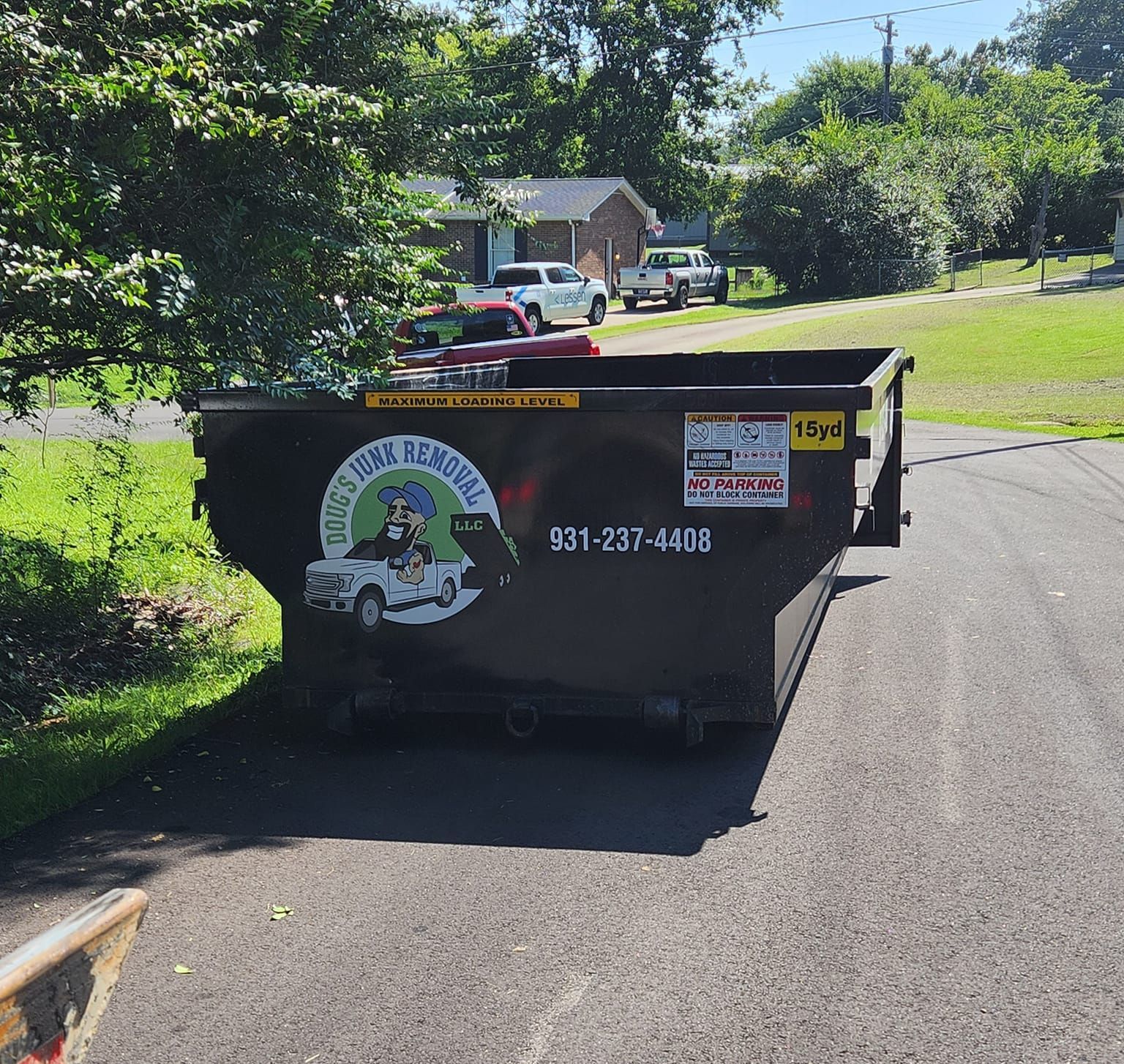 A black dumpster in a driveway, with a logo and phone number, in a suburban setting.