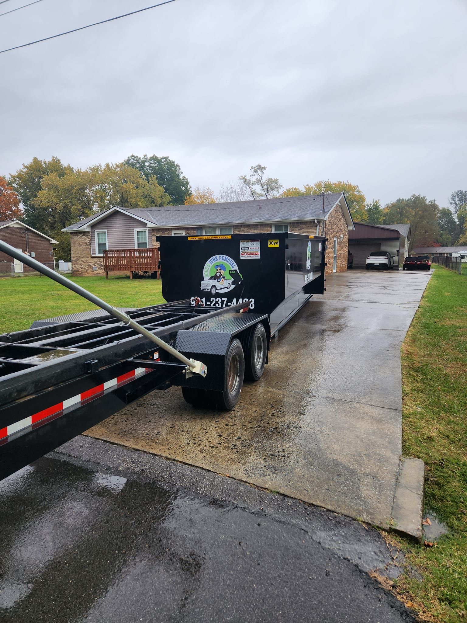 A black trailer with a waste container parked on a driveway in front of a house on a cloudy day.
