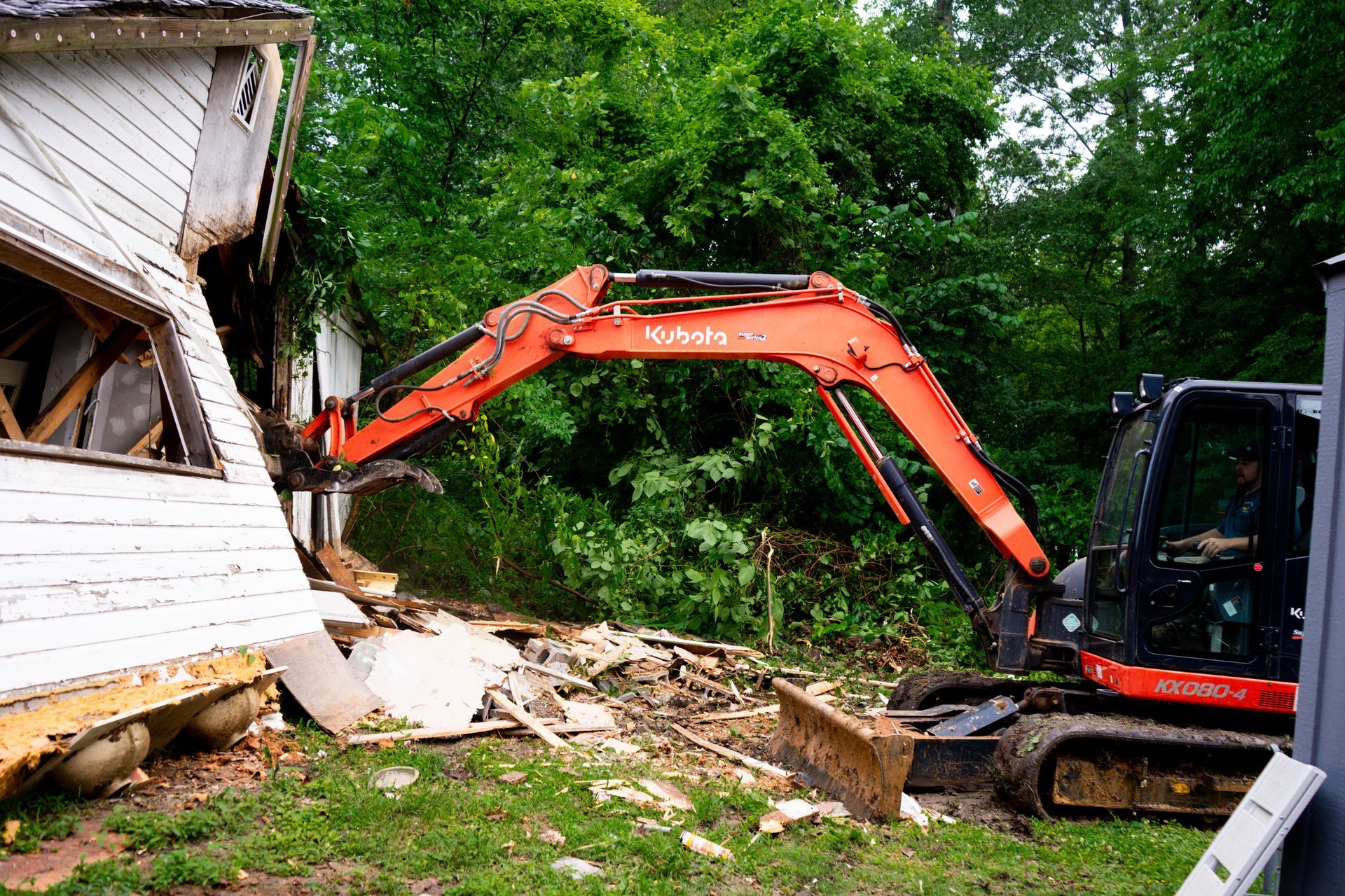 A Kubota excavator demolishes a house, orange arm and claw against white siding, green trees in the background.