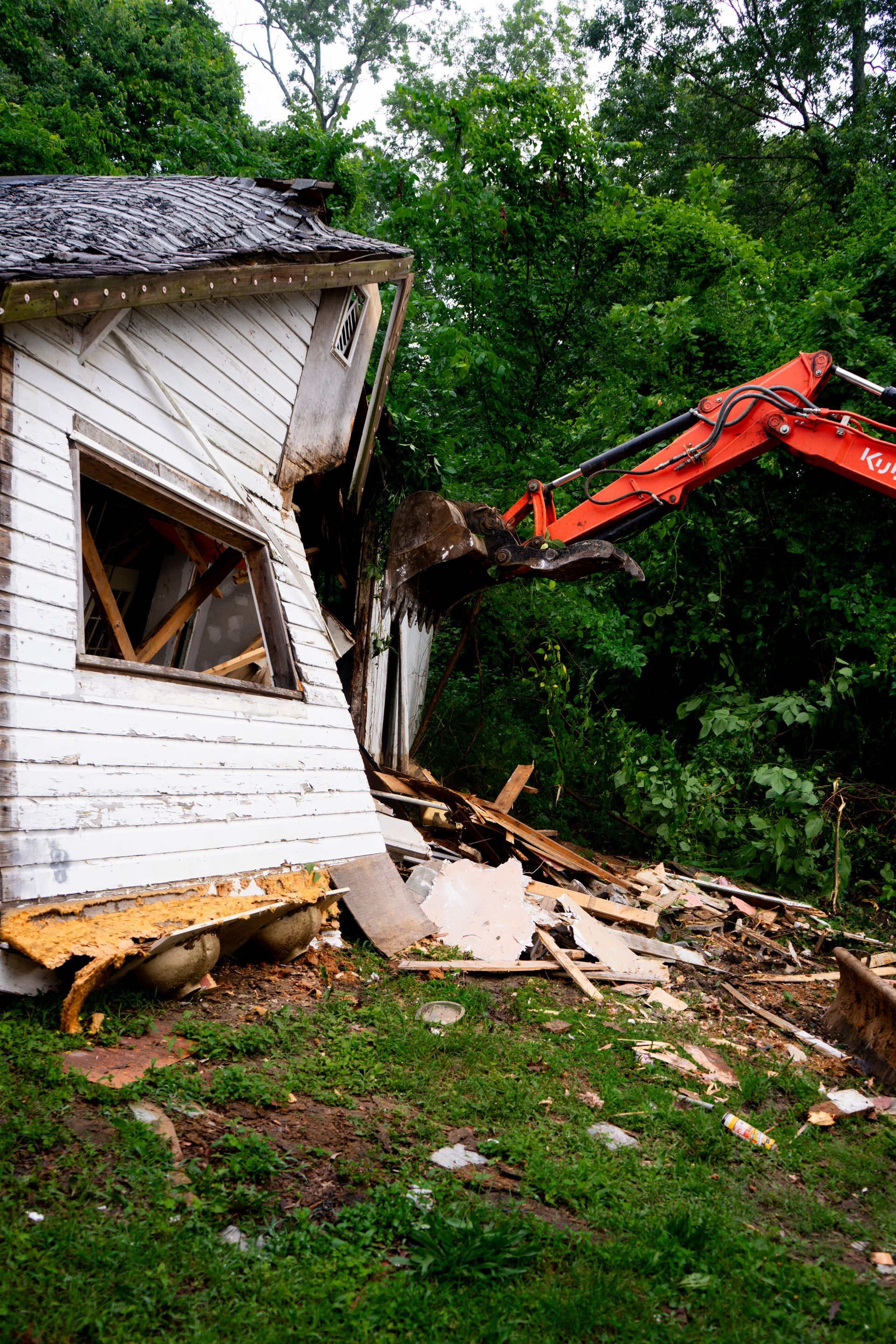 A small, white house being demolished by a red excavator in a wooded area.