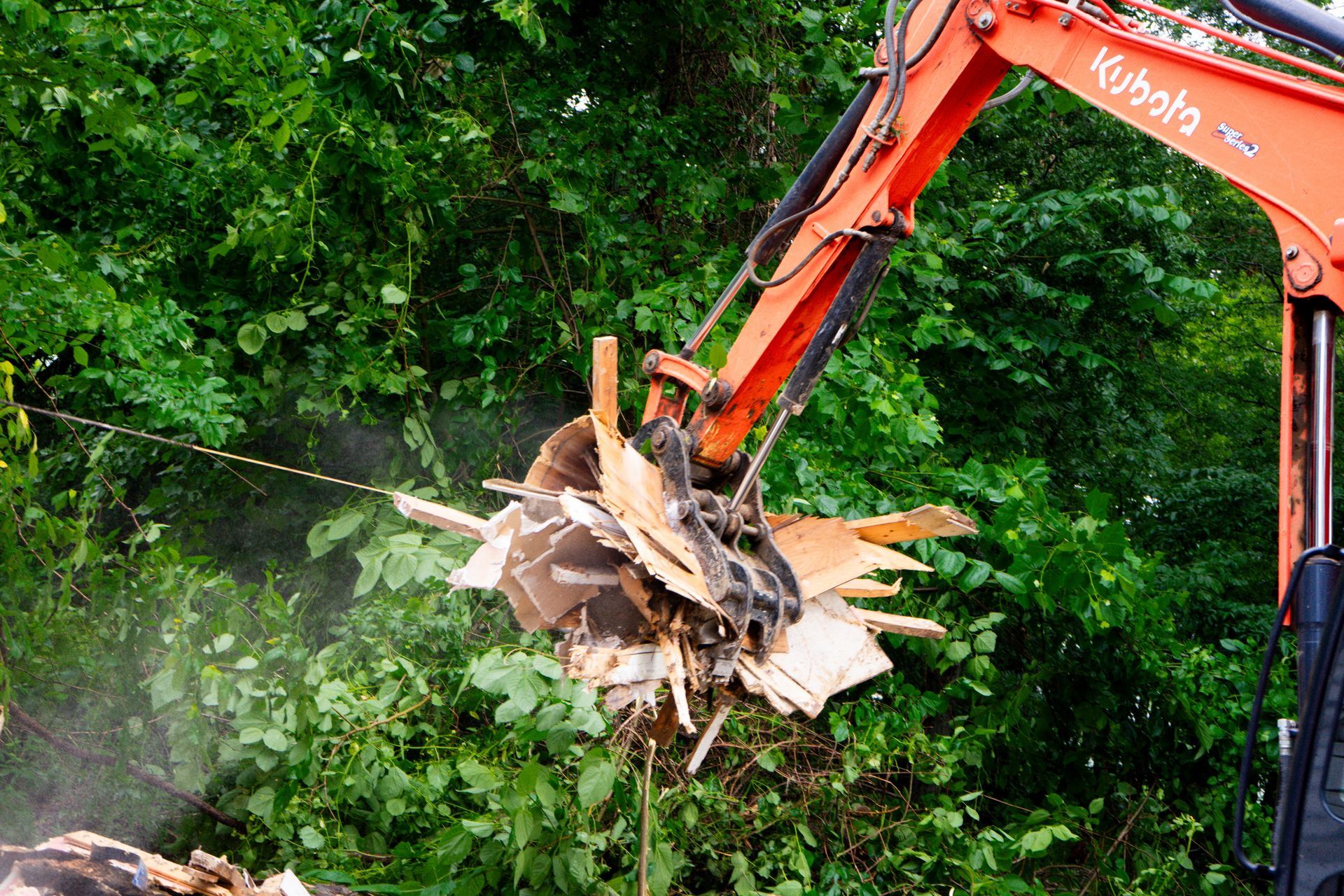 Orange excavator arm grasping debris against a backdrop of green foliage.