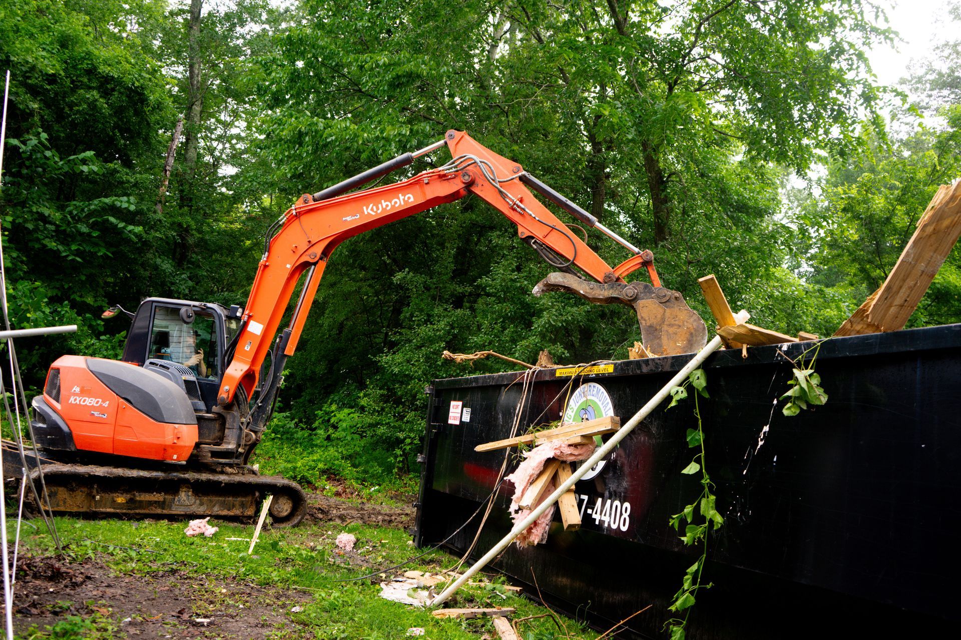 Orange excavator loading debris into a black dumpster in a wooded area.