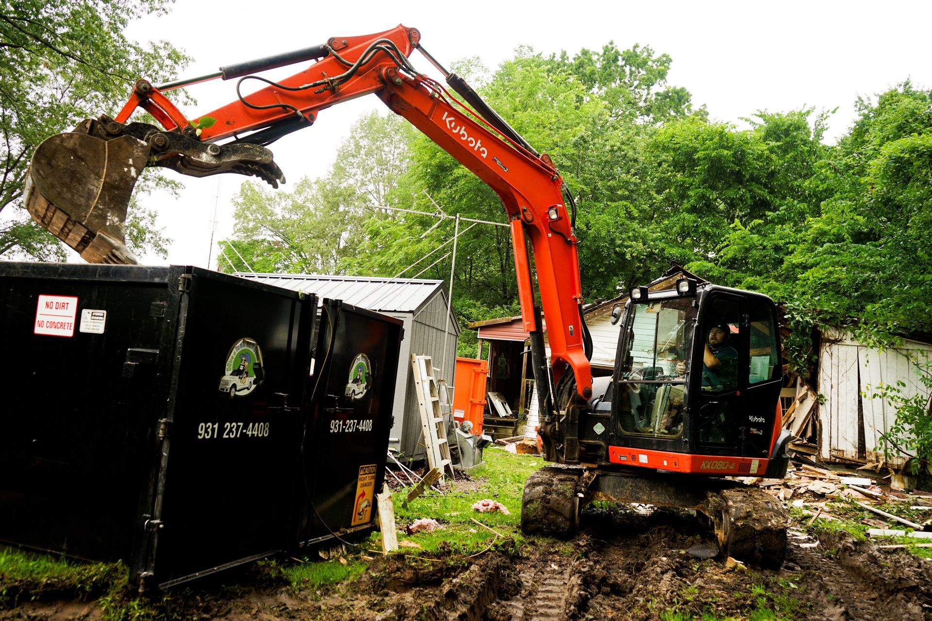 Orange excavator loading debris into a black dumpster in a wooded area.