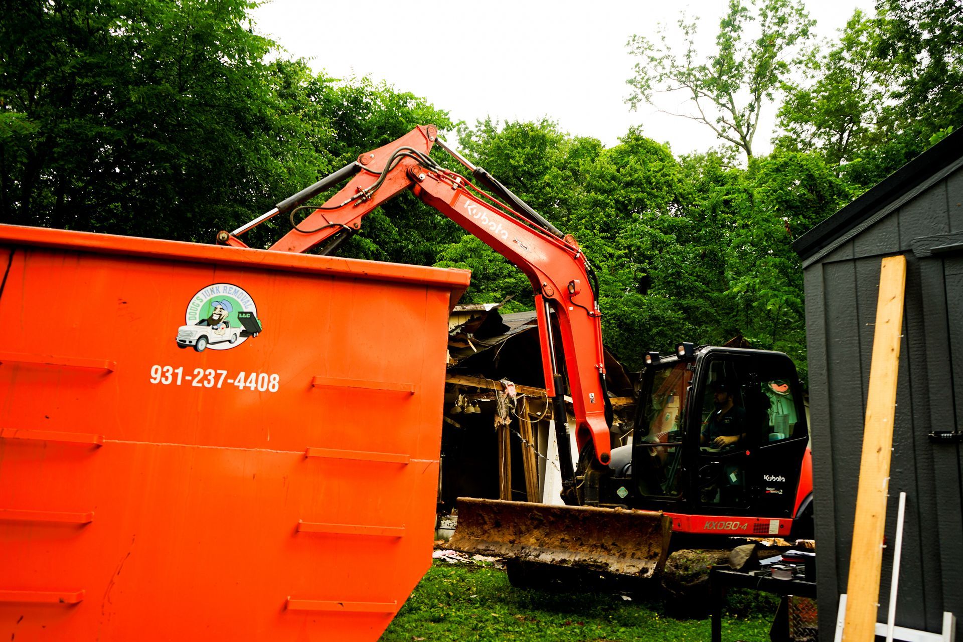 An orange excavator loading debris into a dumpster. Trees and a shed are in the background.