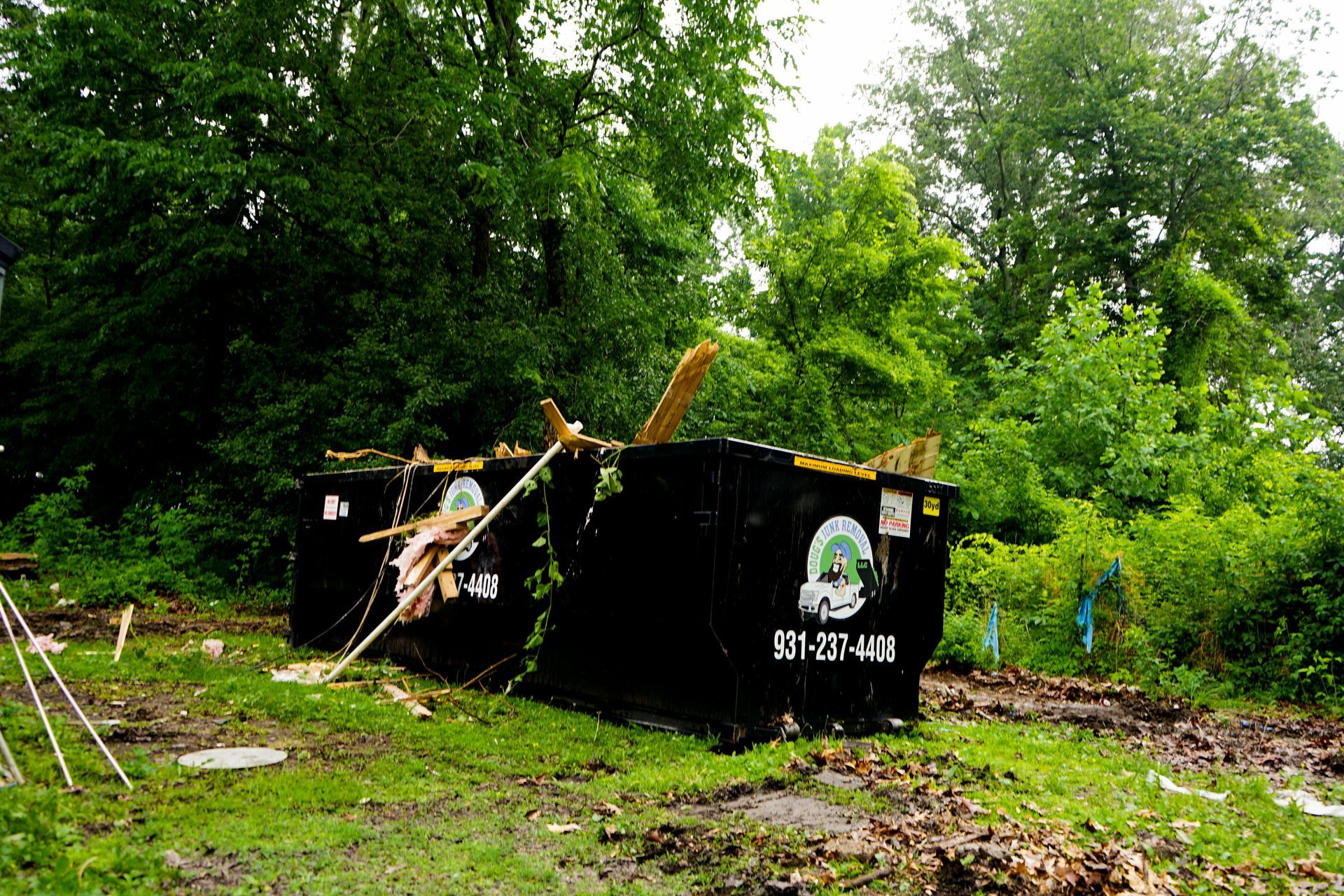 Two black dumpsters overflowing with debris, muddy ground, green foliage background.