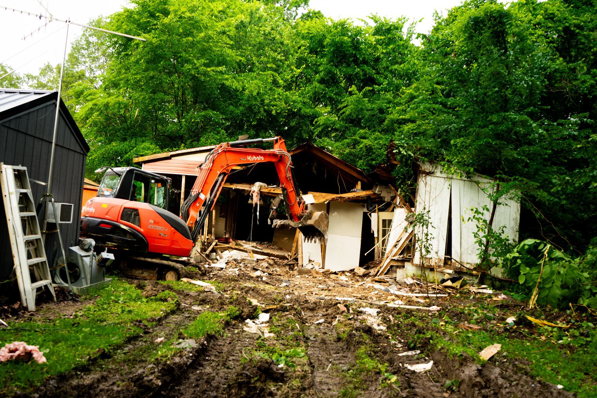 An excavator demolishes a small, white building in a muddy, overgrown yard, surrounded by green trees.