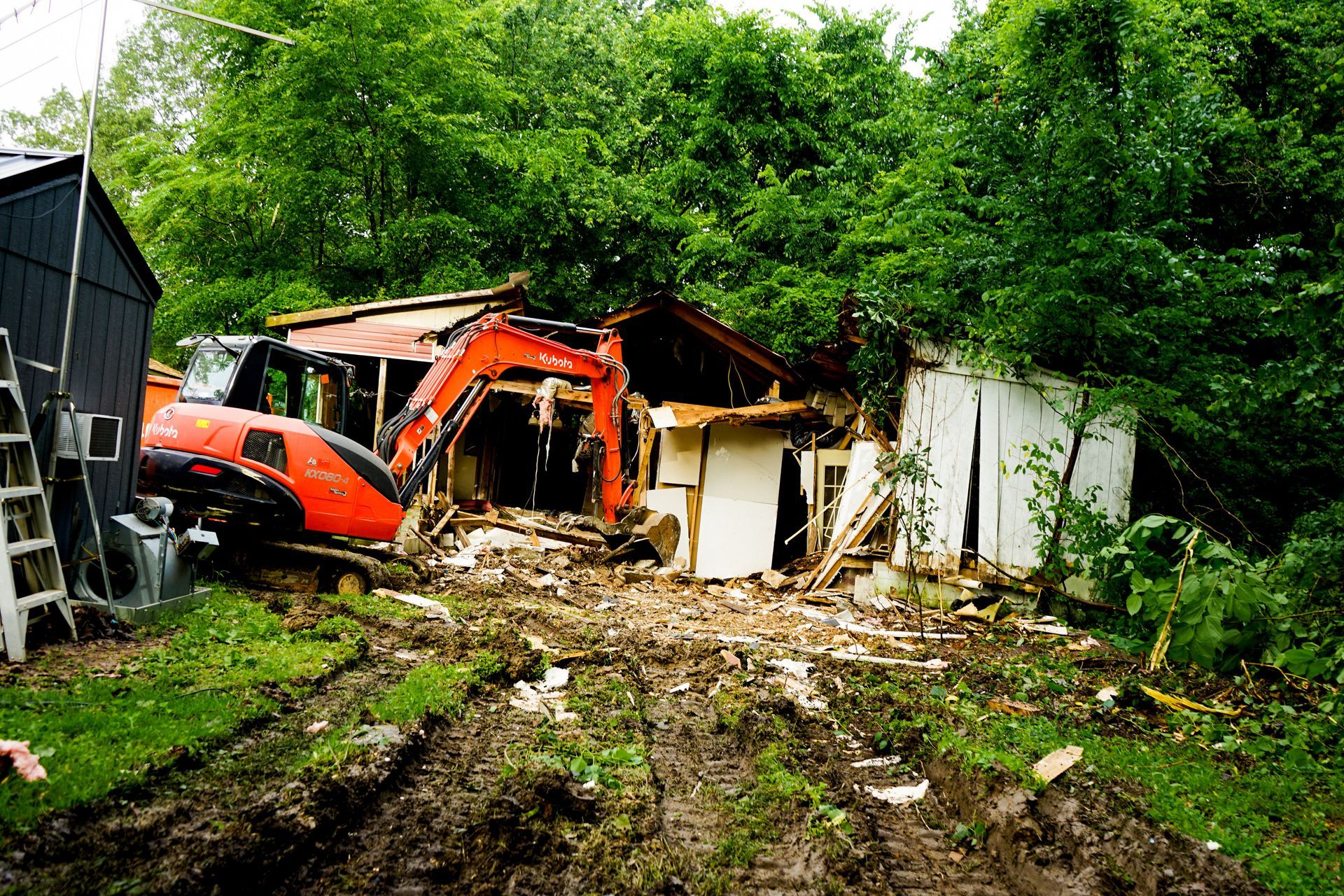Orange excavator demolishing a white and brown house amidst muddy ground and greenery.