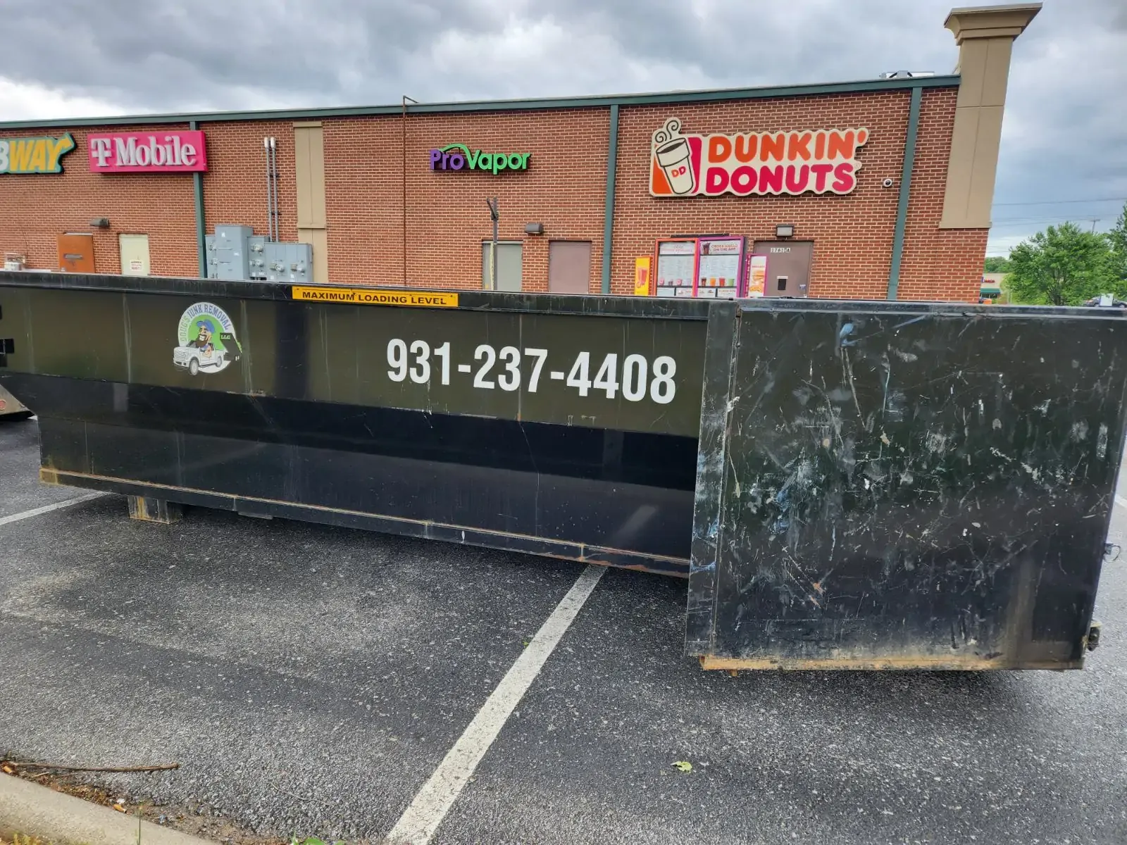 Black dumpster in a parking lot in front of a shopping center, Dunkin' Donuts visible