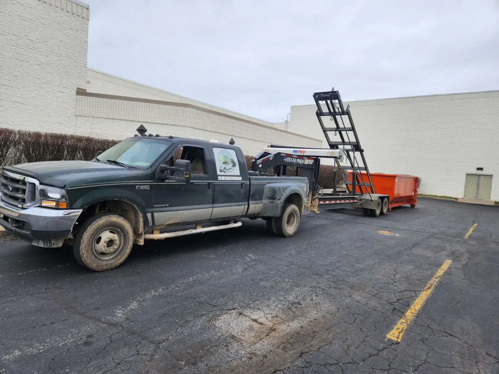 Dark green truck towing orange trailer with lift in front of a white building.
