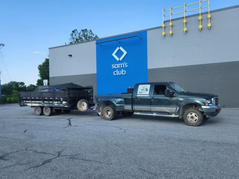 Truck towing a trailer filled with black material in front of a Sam's Club building.